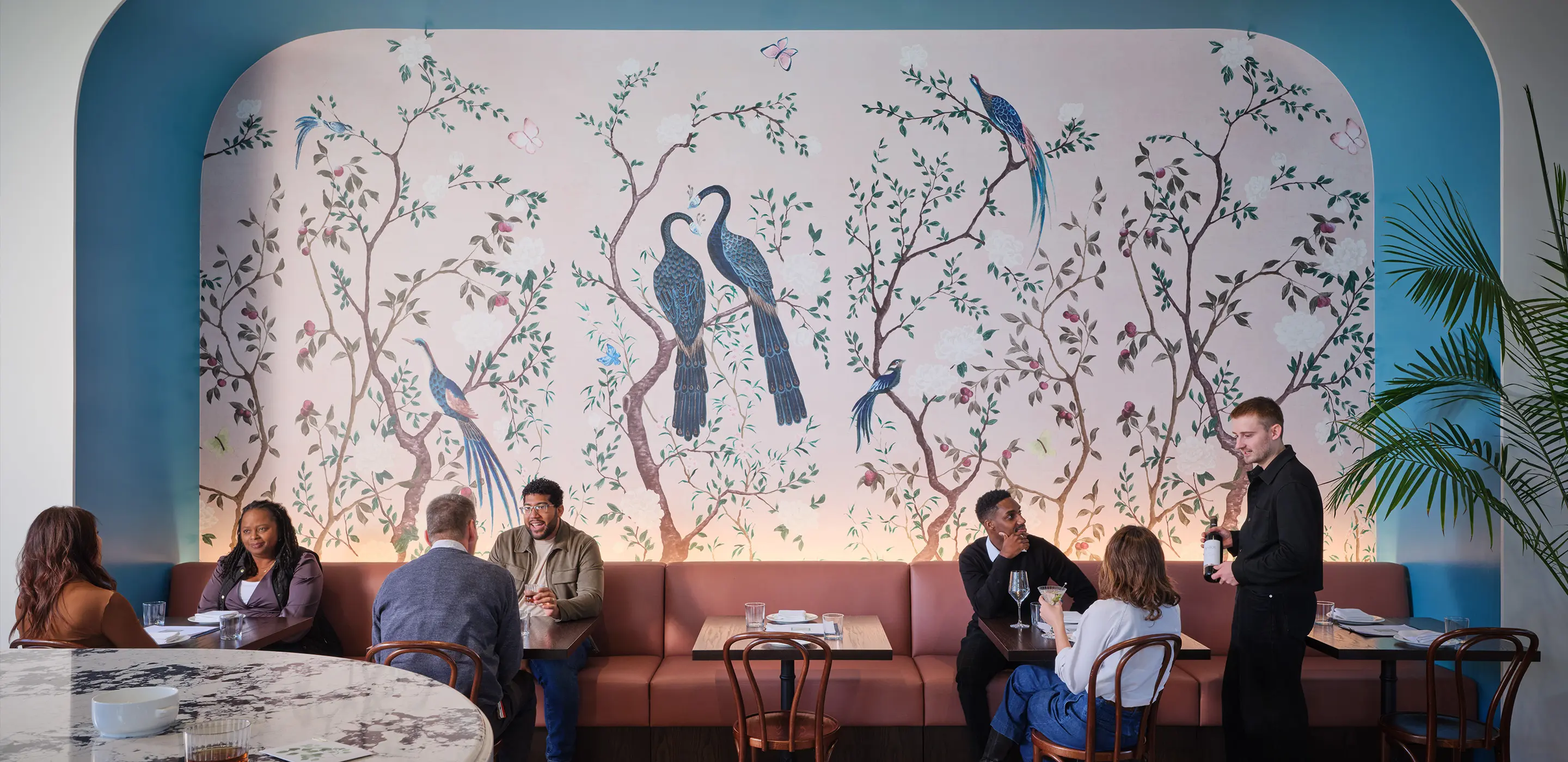 Elegant restaurant interior with floral mural, patrons dining, and waiter serving wine. Cozy ambiance with artistic decor.