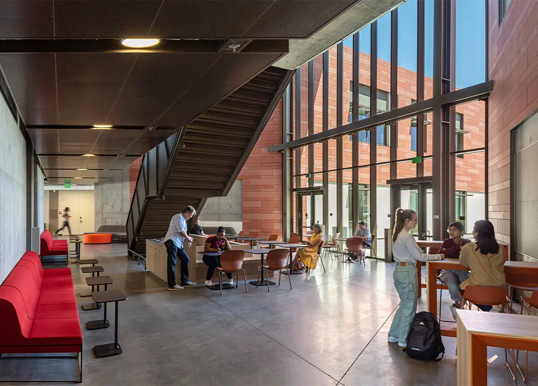 Modern university common area with students studying and socializing under large windows and staircase.