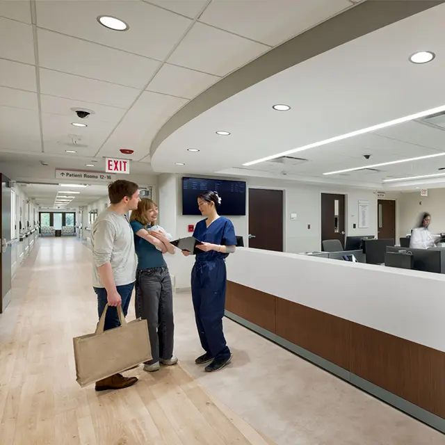 Nurse assisting young couple in modern hospital corridor near reception desk.