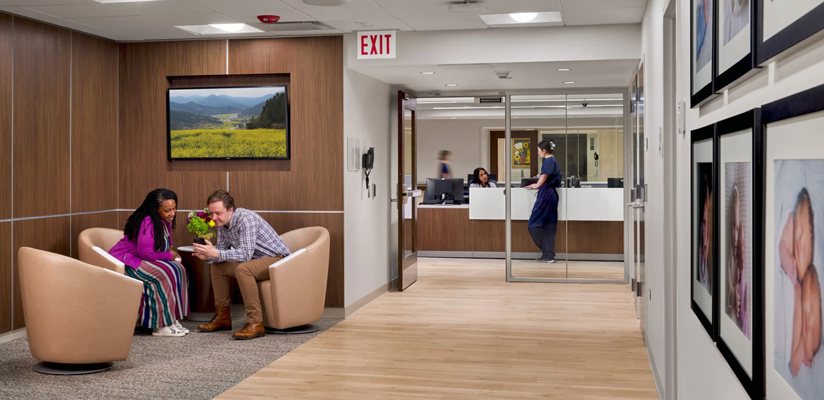 Hospital waiting area with two people seated, vibrant artwork, and medical staff at reception in the background.