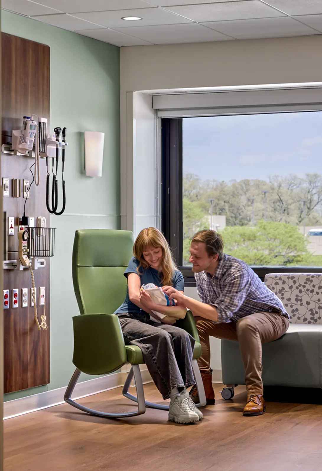 Parents holding their newborn in a hospital room, sitting near medical equipment and large window with greenery outside.