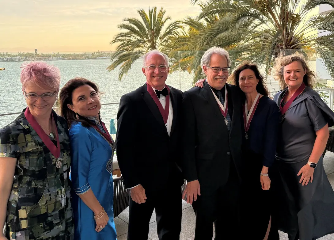 Group of six people in formal attire with medals, standing by waterfront, palm trees in the background.
