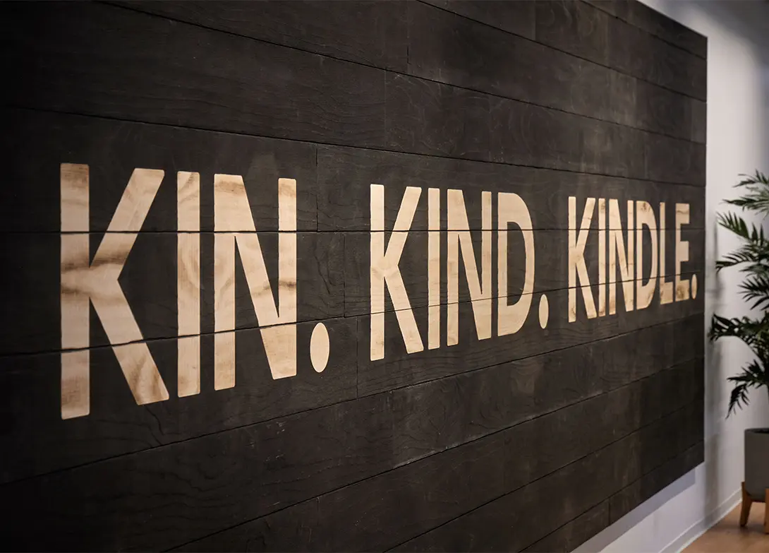 Wall with motivational words KIN. KIND. KINDLE. in bold letters on a dark wooden backdrop.