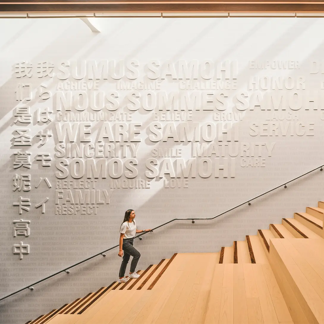 Person walking up modern wooden stairs with WE ARE SAMOHI and multilingual text on wall in bright, airy space.