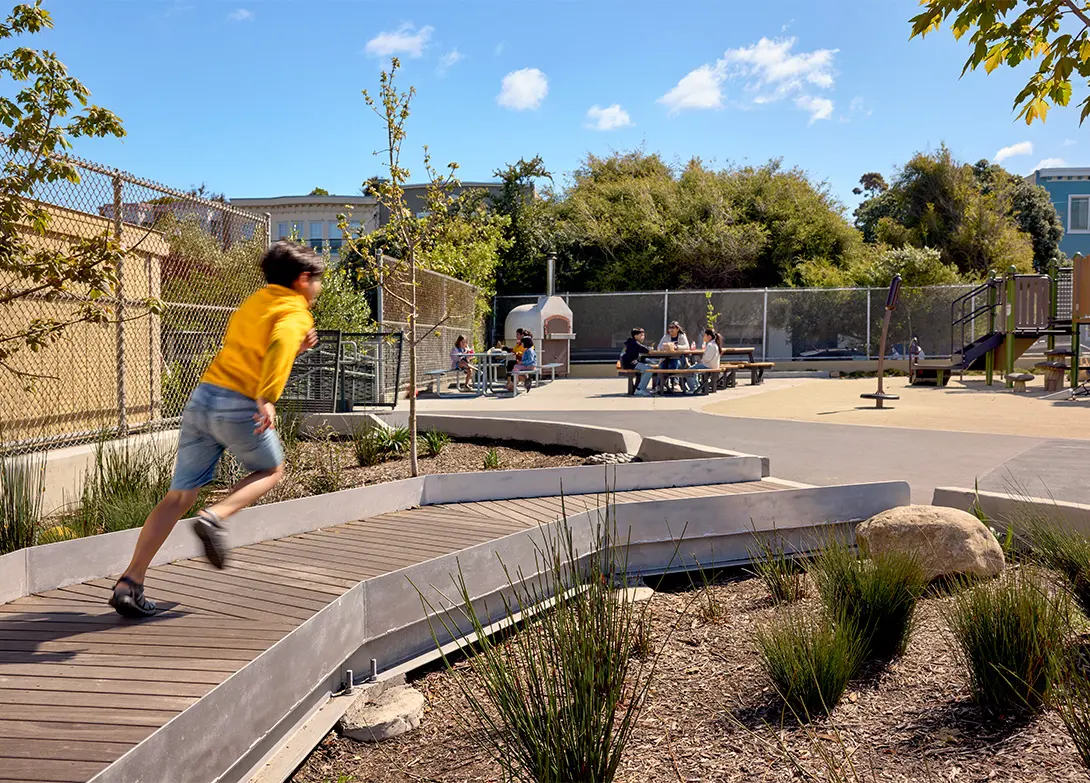 Child running on a playground with tables and trees under a blue sky.