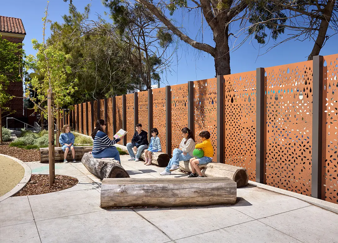 Outdoor learning session with kids and teacher sitting on logs near decorative fence under sunny blue sky.