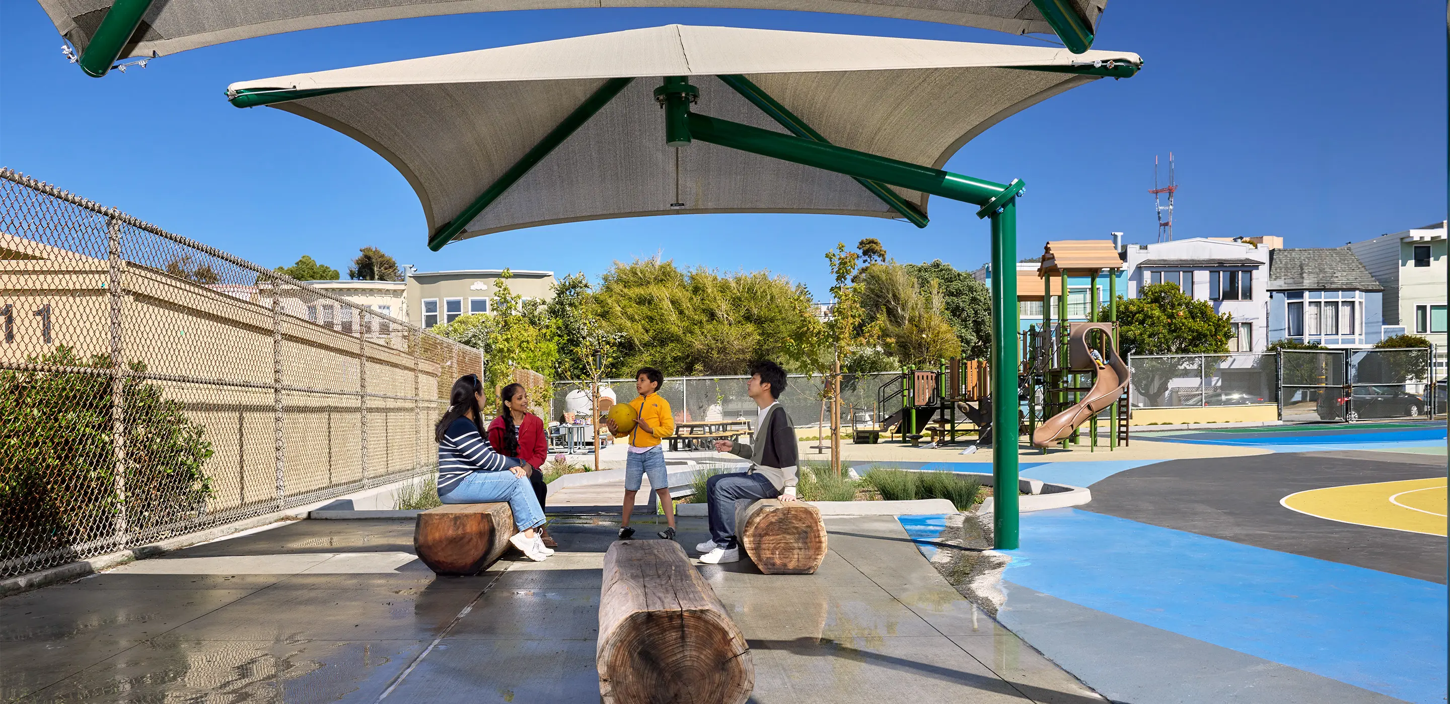 Group of people enjoying a sunny day under a playground canopy with benches and slides nearby.