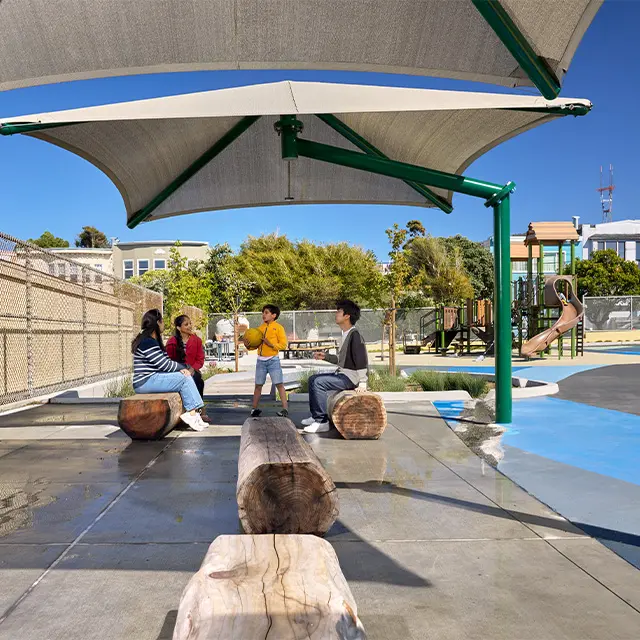 Family enjoying a sunny day at a playground with shade canopy and play structures in the background.