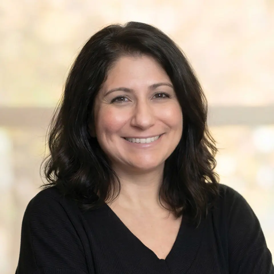 Smiling woman with dark hair, wearing a black shirt, in front of a blurred background indoors.