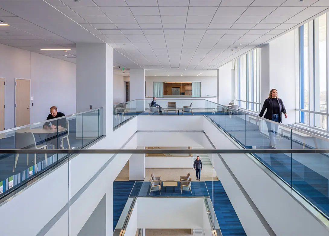 Modern office interior with people working and walking, featuring open spaces and glass railings for natural light.