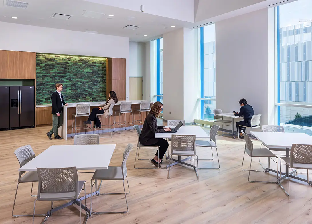 Modern office break room with people working and relaxing, featuring large windows and stylish furniture.