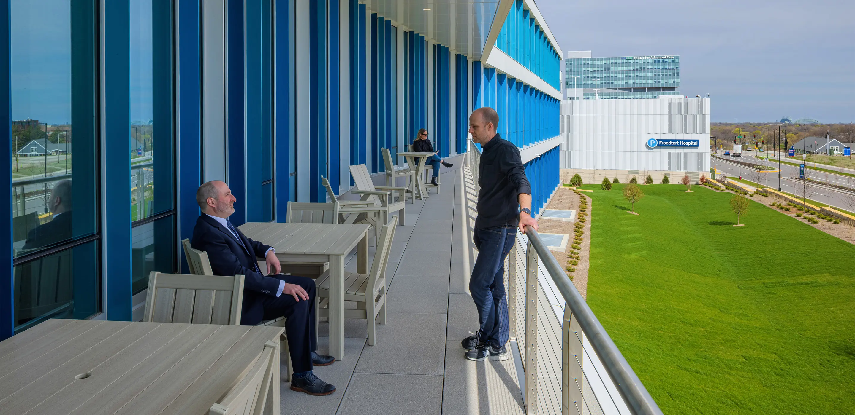 People conversing on balcony with view of Froedtert Hospital building and green lawn.