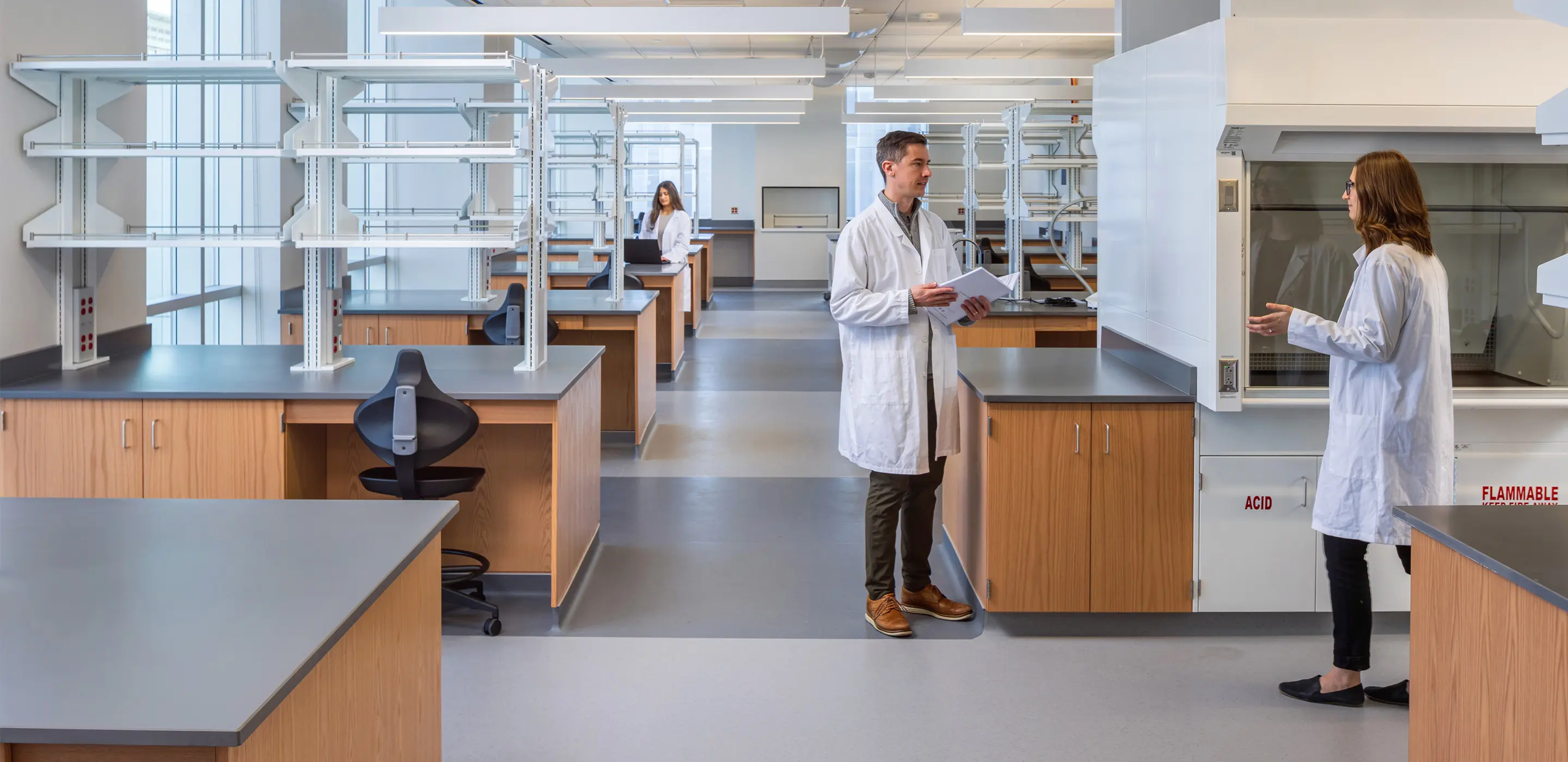 Scientists in lab coats discussing in a spacious, modern laboratory with wooden cabinets and ample shelving.