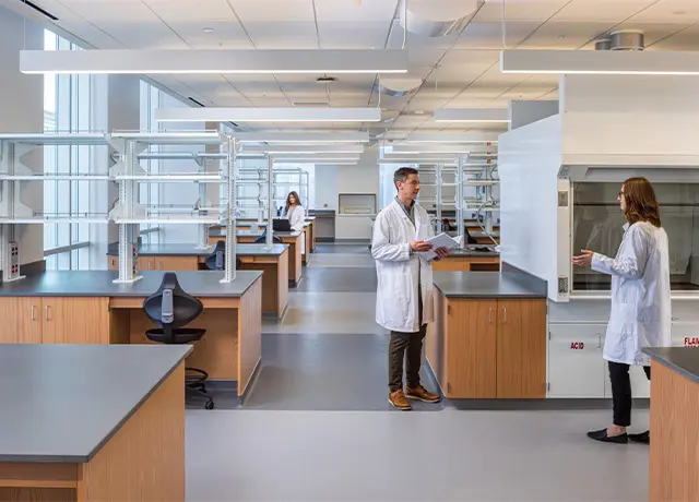 Scientists in lab coats discuss inside a modern laboratory with workstations and lab equipment.
