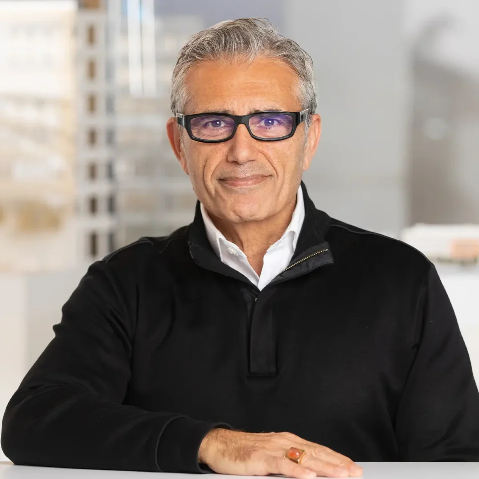 Elderly man with glasses and black sweater smiling in a bright office setting.