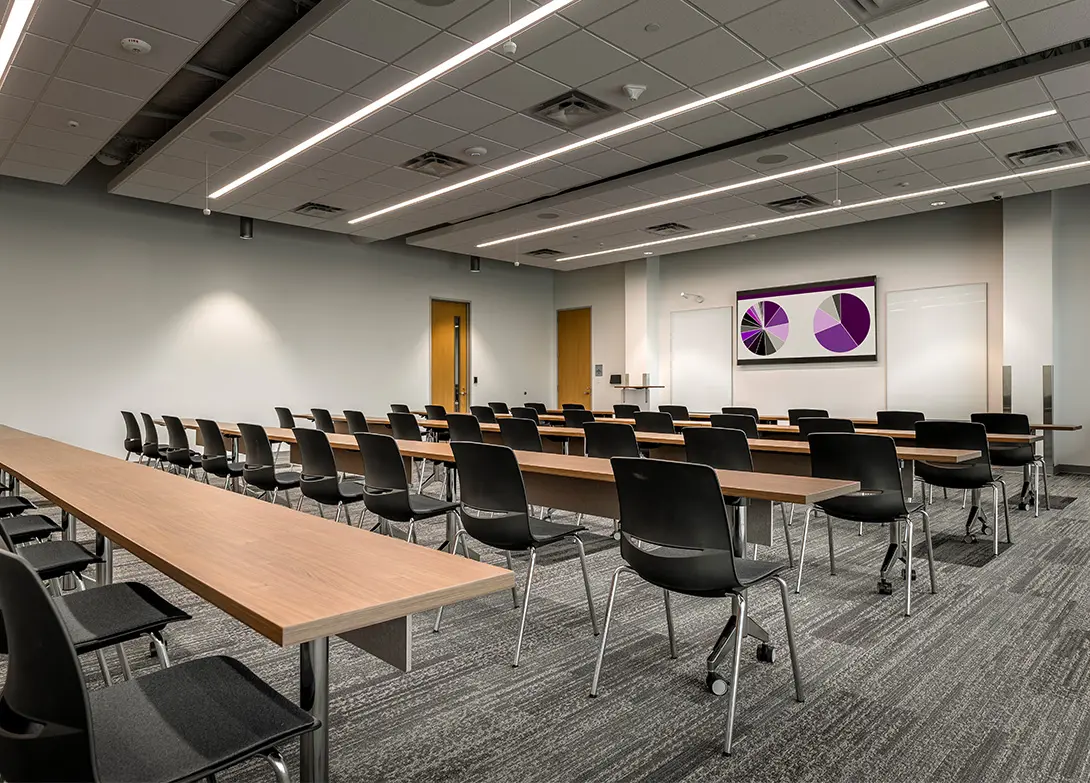 Modern conference room with rows of empty chairs and tables, featuring a large screen displaying colorful charts.