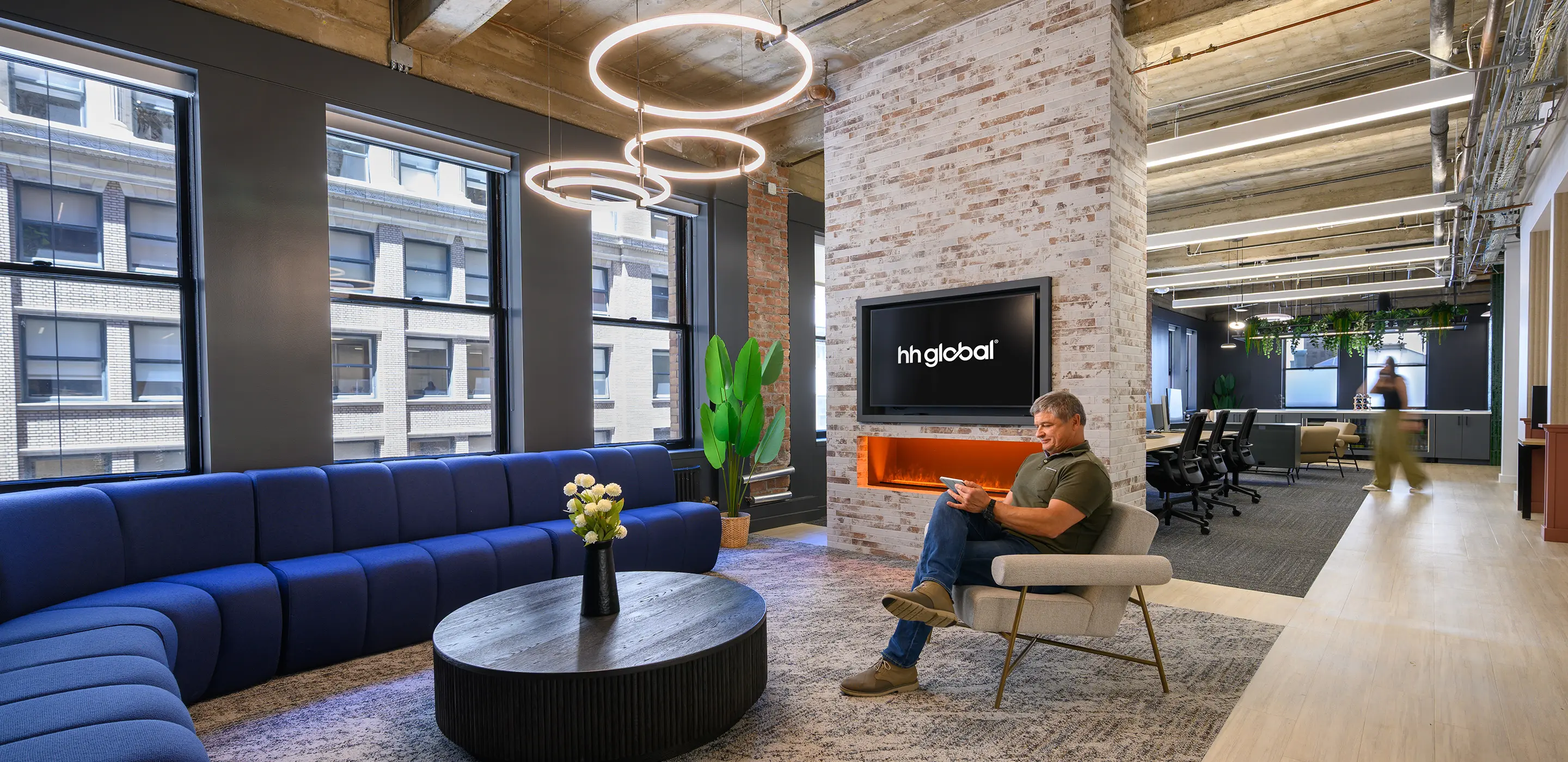 Modern office lounge with blue sofa, round table, and man reading near a plant and TV. Bright, contemporary workspace.