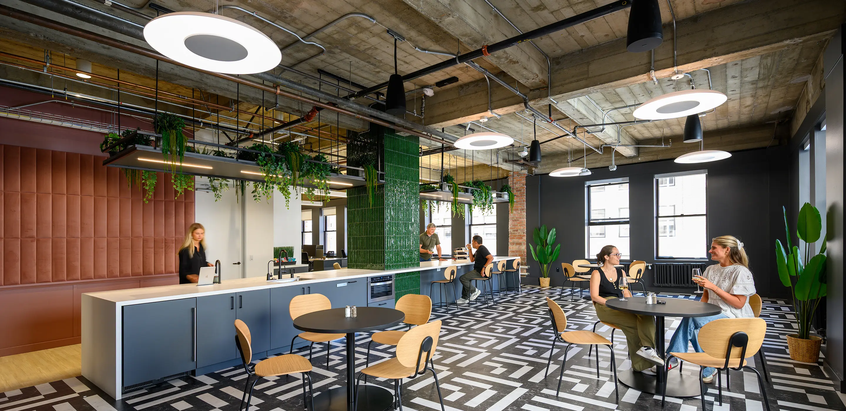 Modern office break area with stylish decor, plants, and people at tables, creating a relaxed atmosphere.