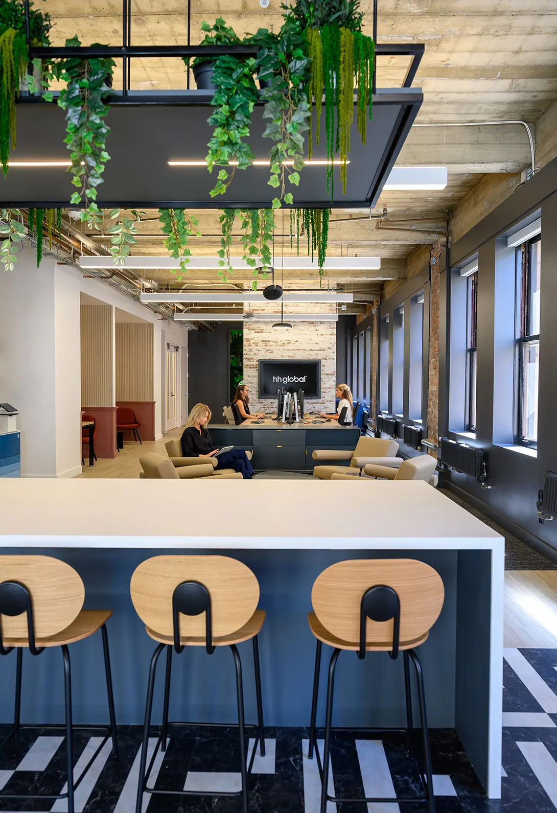 Modern office space with lush green plants, stylish chairs, and people working at computers and relaxing in seating area.
