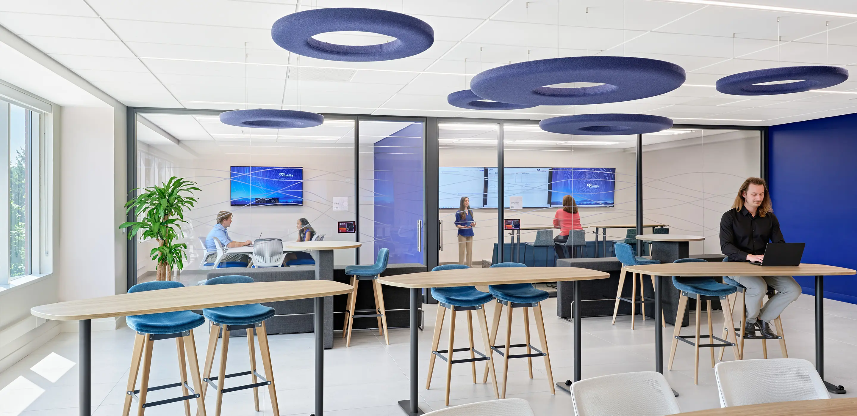 Modern office with collaborative workspaces, blue accents, and people engaged in meetings and laptop work.